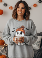 Woman wearing a gray sweatshirt with a ghost design holding a mug in a Halloween-themed room.