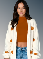 Woman wearing a white cardigan with pumpkin patterns, burnt orange top, and blue jeans on a gray background