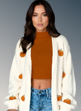 Woman wearing a white cardigan with pumpkin patterns, burnt orange top, and blue jeans on a gray background