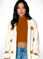 Woman wearing a white cardigan with orange pumpkin patterns, burnt orange top, and blue jeans on a white background